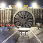 Night-time construction site showing a large TBM tunnel boring machine face, with crowd of workers in safety vests watching behind barriers and banners overhead