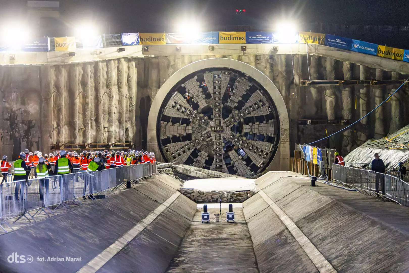 Night-time construction site showing a large TBM tunnel boring machine face, with crowd of workers in safety vests watching behind barriers and banners overhead