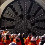 Construction crew in orange safety gear gathered around a massive tunnel-boring machine cutter head inside a tunnel