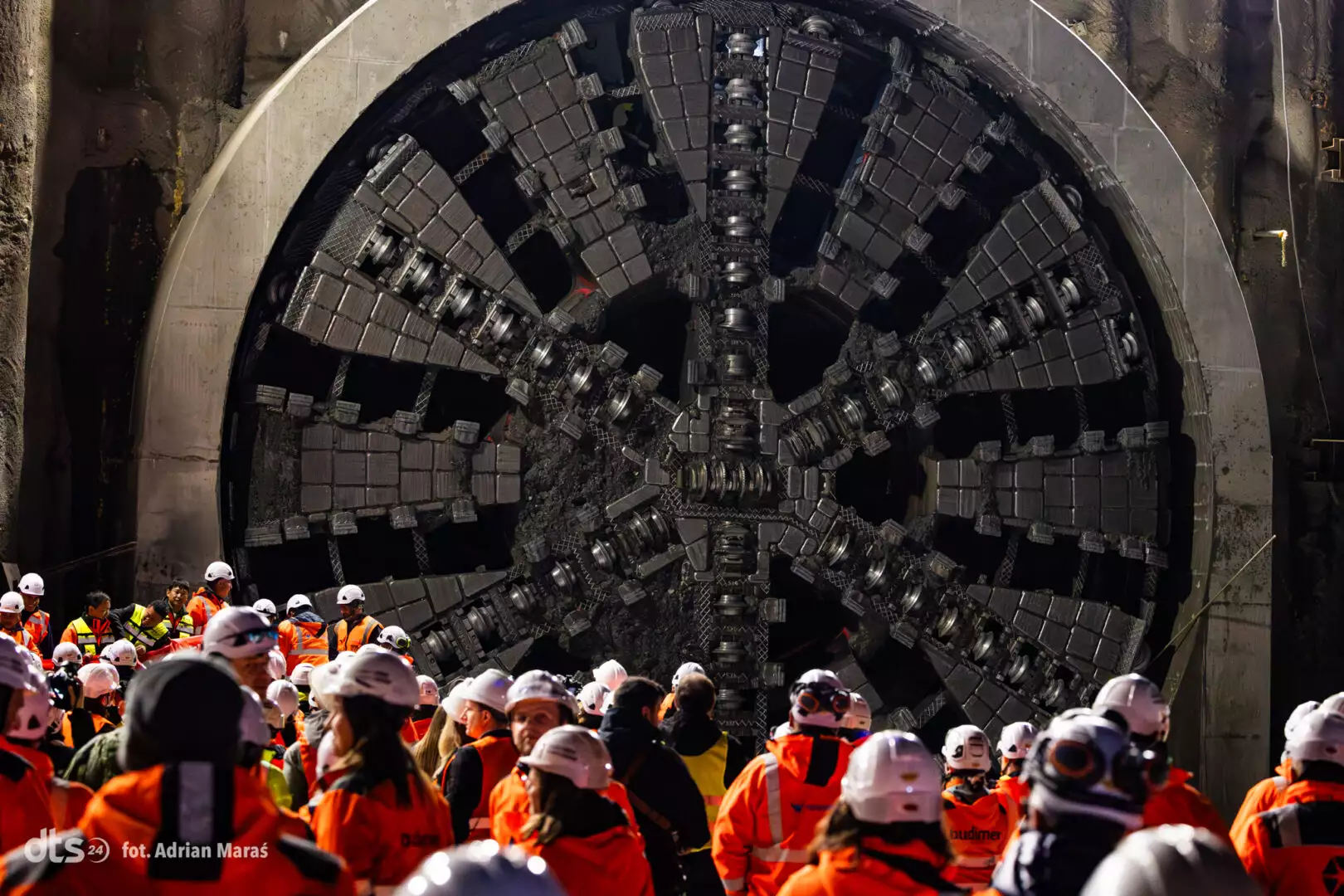 Construction crew in orange safety gear gathered around a massive tunnel-boring machine cutter head inside a tunnel