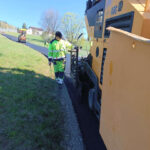 Road construction scene: worker in neon safety gear guiding asphalt being laid by a large yellow paver roller.