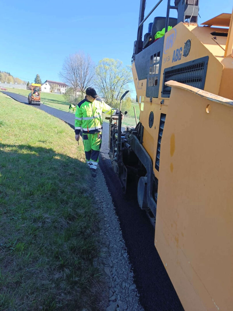 Road construction scene: worker in neon safety gear guiding asphalt being laid by a large yellow paver roller.