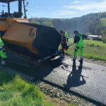Road construction crew in neon gear operating a paving machine laying fresh asphalt on a rural road.