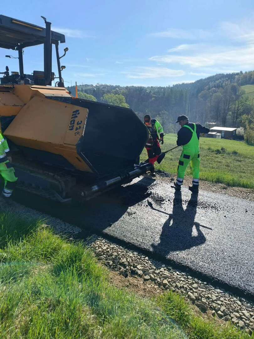 Road construction crew in neon gear operating a paving machine laying fresh asphalt on a rural road.