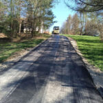 Paved road under construction with a yellow paving truck ahead; trees line the sides on a sunny day.