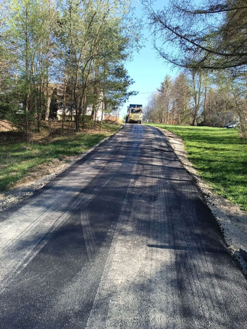 Paved road under construction with a yellow paving truck ahead; trees line the sides on a sunny day.