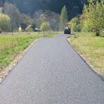 Freshly paved rural road curves toward a road roller in the distance, with a small house and trees on the left.