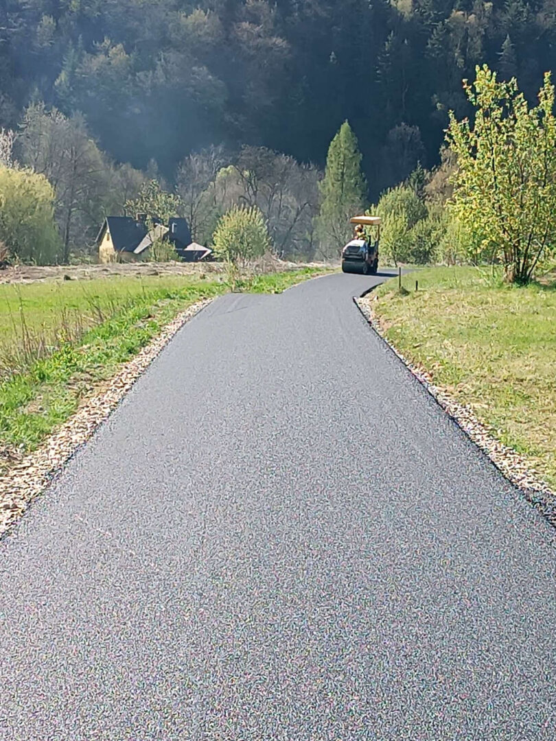 Freshly paved rural road curves toward a road roller in the distance, with a small house and trees on the left.