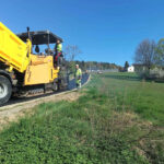 Yellow asphalt paver in operation on a rural road, two workers in high-visibility vests nearby under a clear blue sky, laying new pavement.