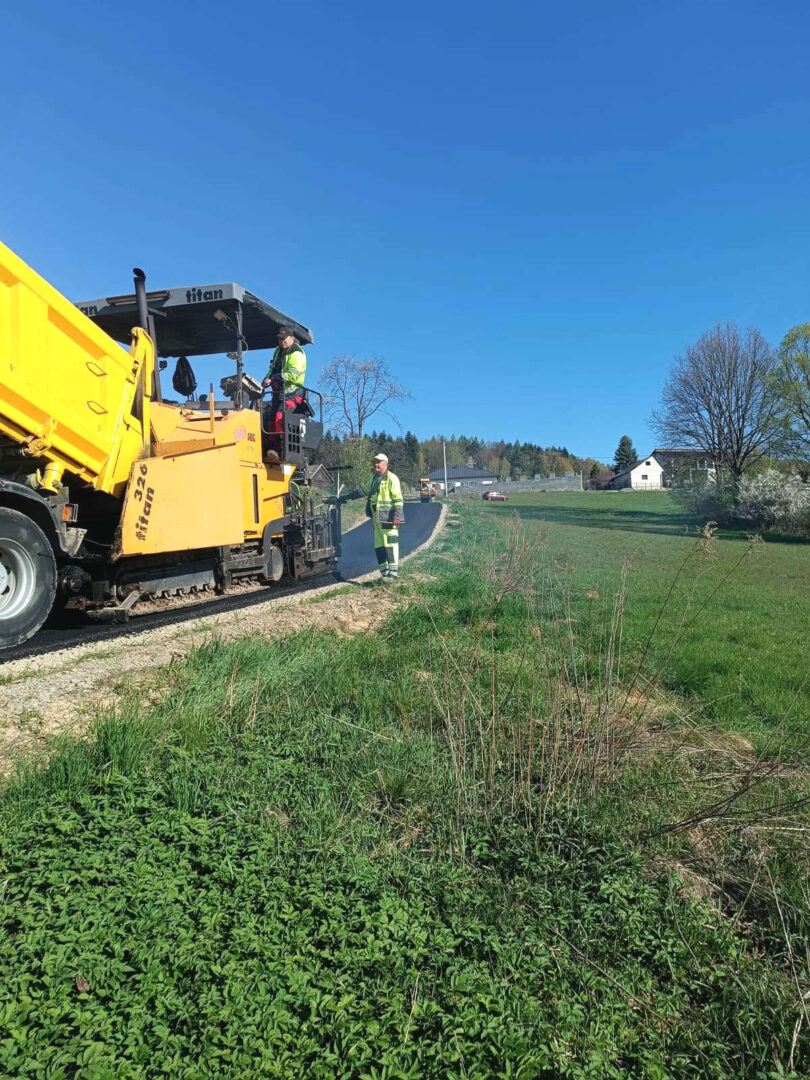 Road construction scene: a large yellow asphalt paver with two workers in high-visibility vests guiding the machine on a rural road.