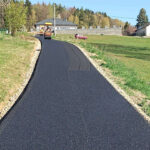 Freshly paved rural road with an asphalt roller on the left, a red car in the distance, and green fields with trees.