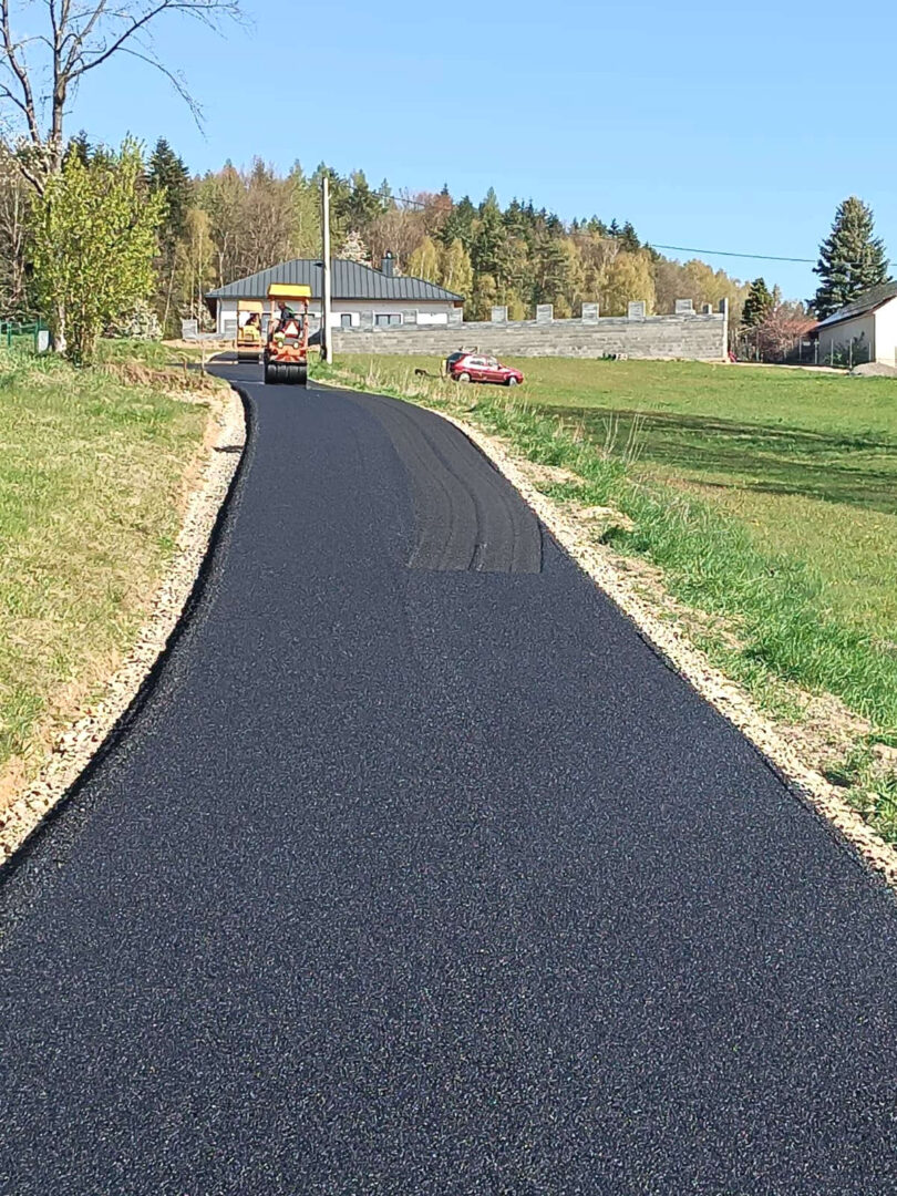 Freshly paved rural road with an asphalt roller on the left, a red car in the distance, and green fields with trees.