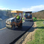 Road construction scene: workers in high-visibility gear lay fresh asphalt with a paving machine and roller on a rural road edge.