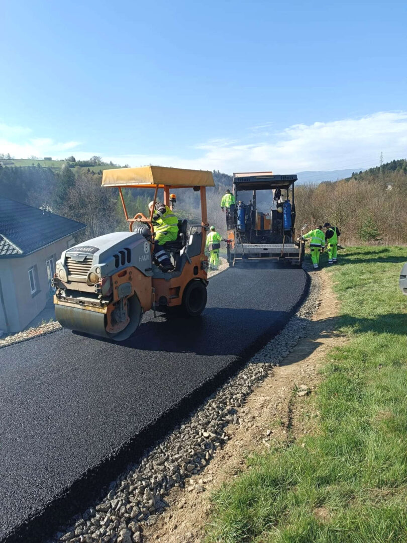 Road construction scene: workers in high-visibility gear lay fresh asphalt with a paving machine and roller on a rural road edge.