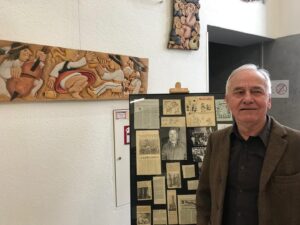 Older man in a brown blazer stands beside a history display board with newspaper clippings and photos in a gallery.