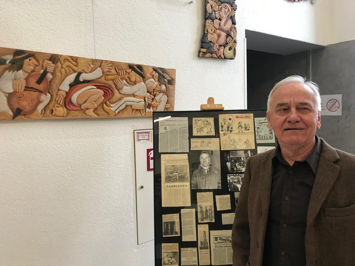 Older man in a brown blazer stands beside a history display board with newspaper clippings and photos in a gallery.