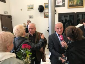 Older man in a suit is interviewed by a reporter with a pink microphone at an indoor social event, paintings on the wall behind them.