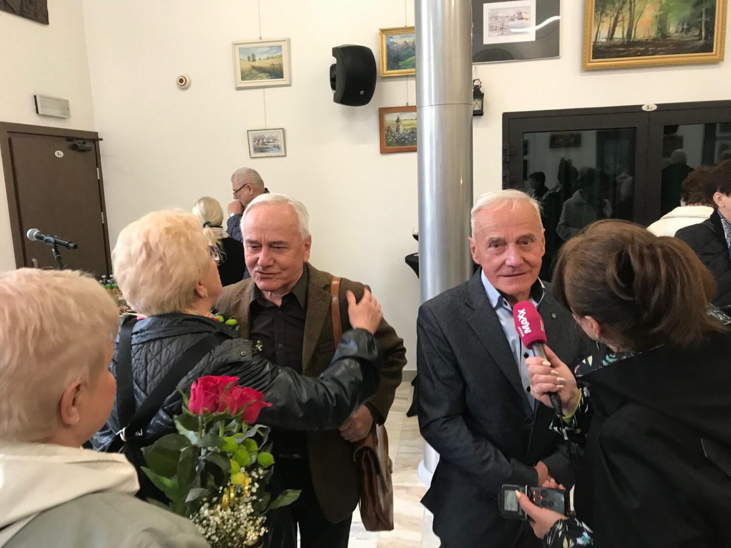 Older man in a suit is interviewed by a reporter with a pink microphone at an indoor social event, paintings on the wall behind them.