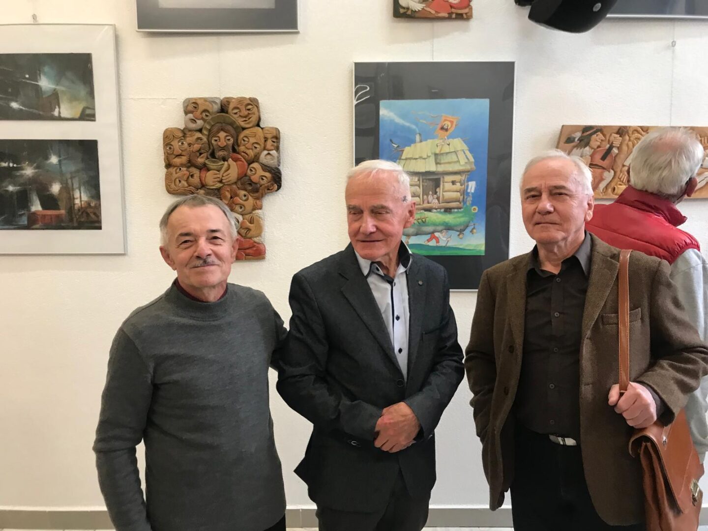 Three older men posing together in an art gallery, smiling at the camera, with framed artworks on the white wall behind them.