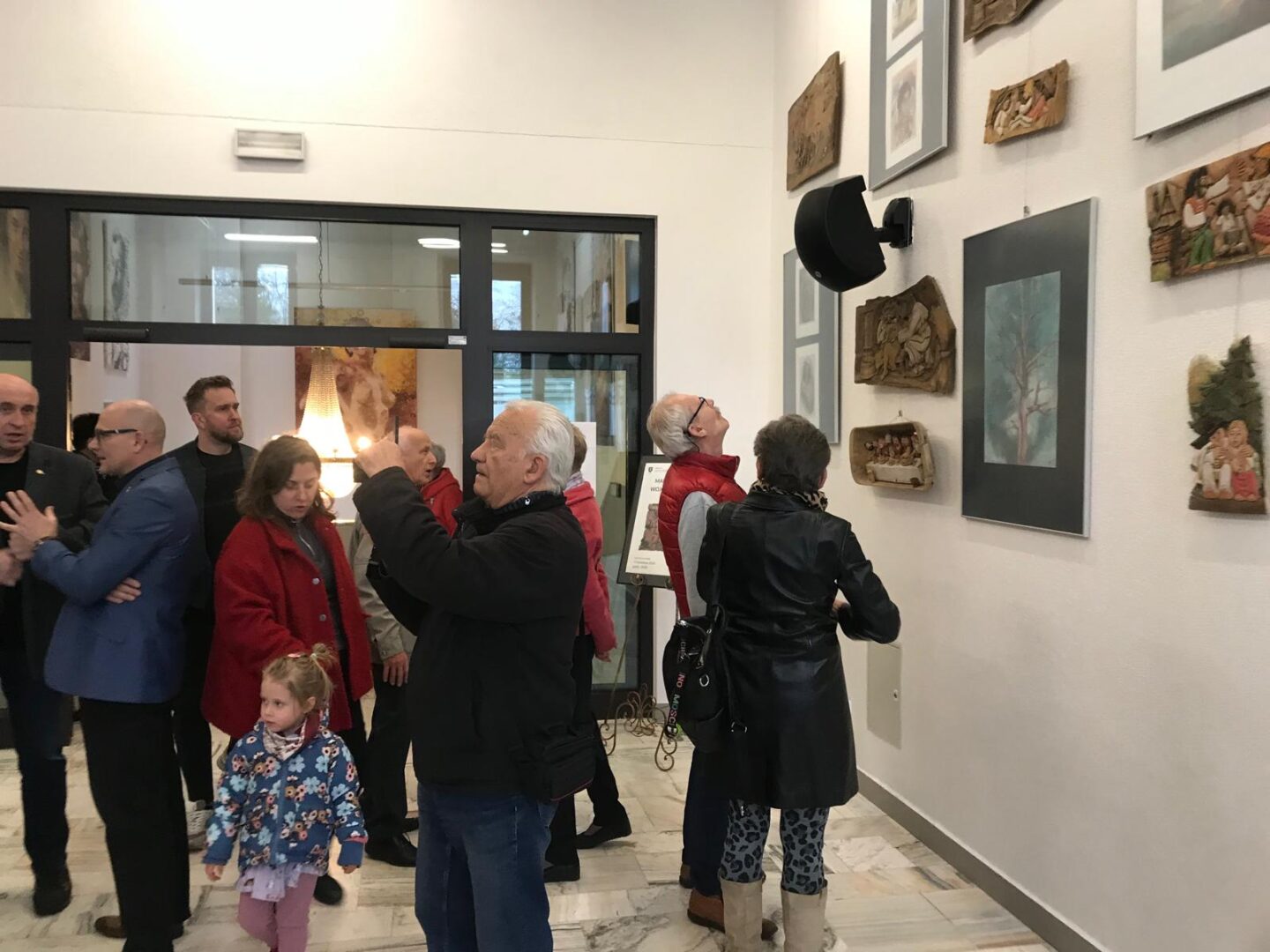 Guests at an art gallery opening gather in a bright hallway, inspecting wall-mounted frames and small sculptural pieces. The group includes adults and a child in a floral jacket foreground on marble floors.