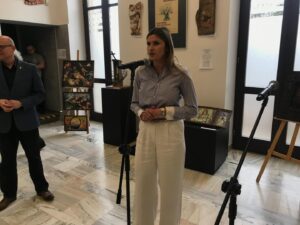 Woman speaker wearing a striped shirt and white wide-leg pants stands at a mic in a gallery, with artwork on display behind her.
