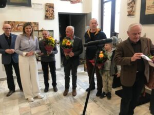 Group of people at an indoor ceremony holding bouquets of flowers, with a microphone in foreground and a woman in white pants center-left smiling.