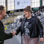 Man wearing a headset and black suit shakes hands with a smiling woman in a leather jacket and leopard-print skirt in a stadium concourse.