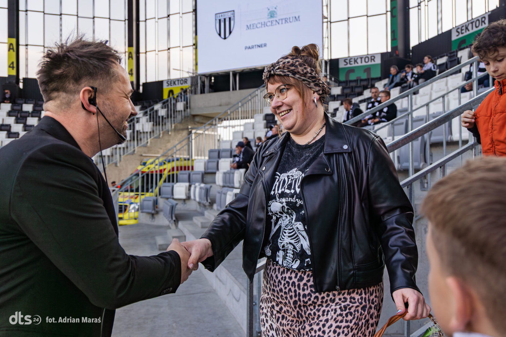 Man wearing a headset and black suit shakes hands with a smiling woman in a leather jacket and leopard-print skirt in a stadium concourse.