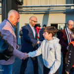 Man in a navy blazer shakes hands with a boy in a white hoodie at an outdoor event, while others chat in the background.