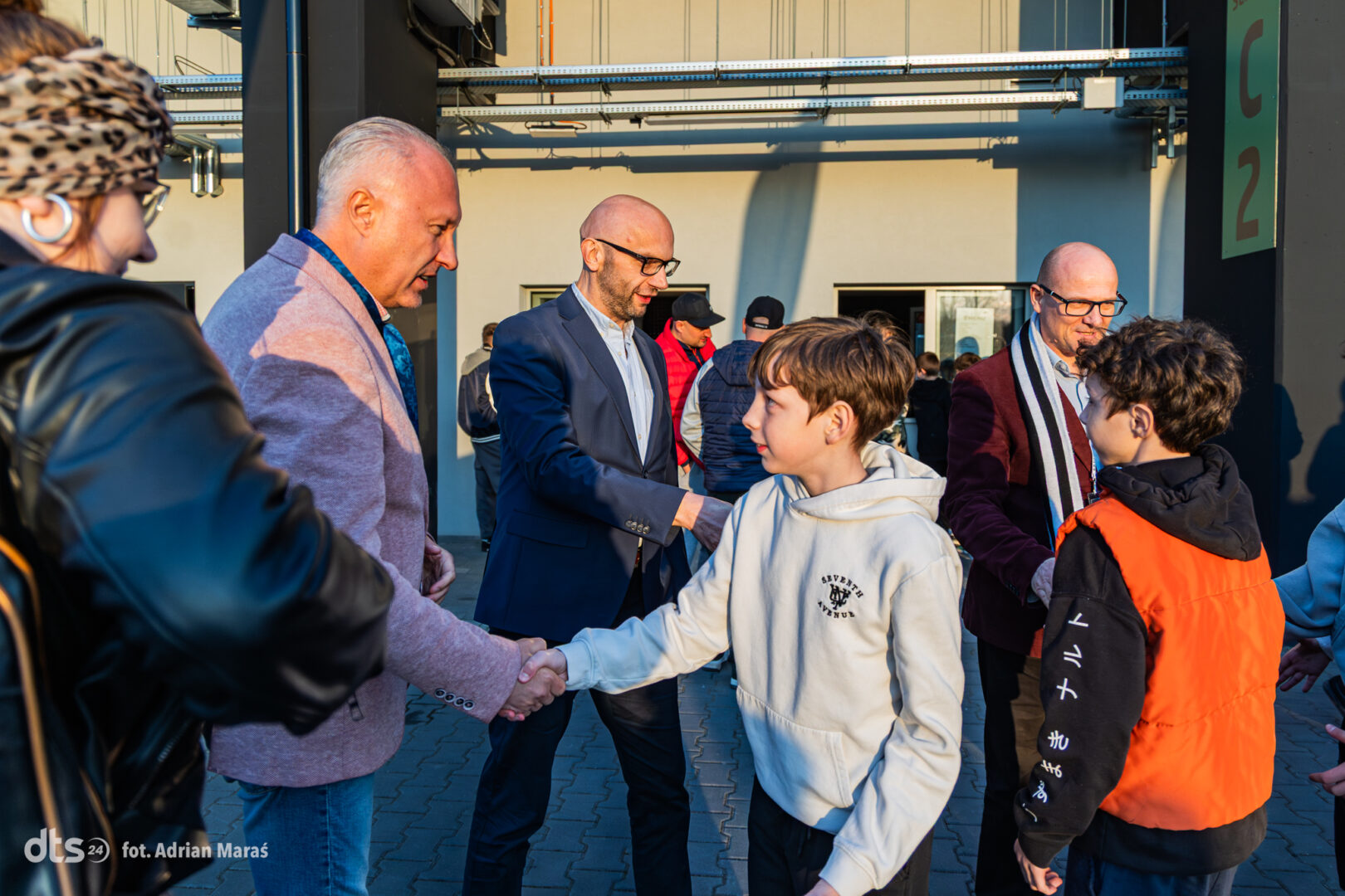 Man in a navy blazer shakes hands with a boy in a white hoodie at an outdoor event, while others chat in the background.