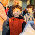 A smiling boy in an orange puffer vest stands in a crowd outdoors, with adults nearby in casual jackets during daylight.