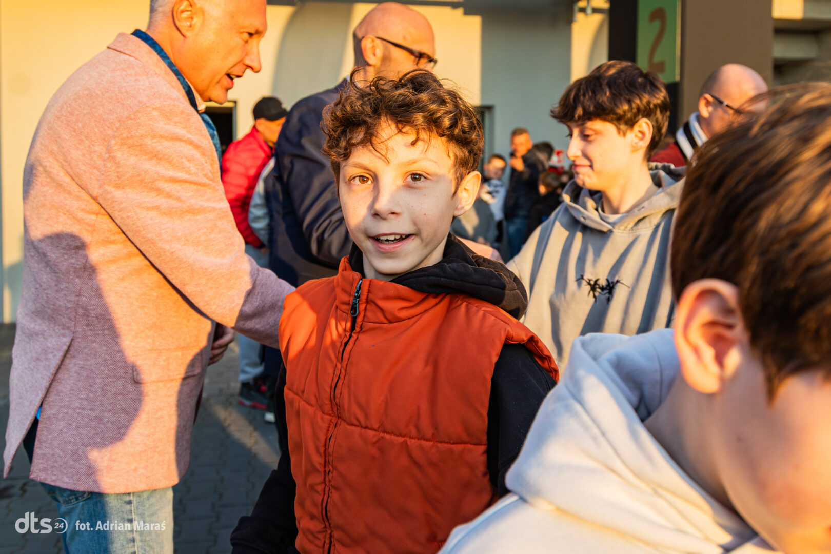 A smiling boy in an orange puffer vest stands in a crowd outdoors, with adults nearby in casual jackets during daylight.