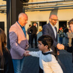 Two men in blazers shake hands with young people at an outdoor event, sunlit courtyard, others socializing in the background