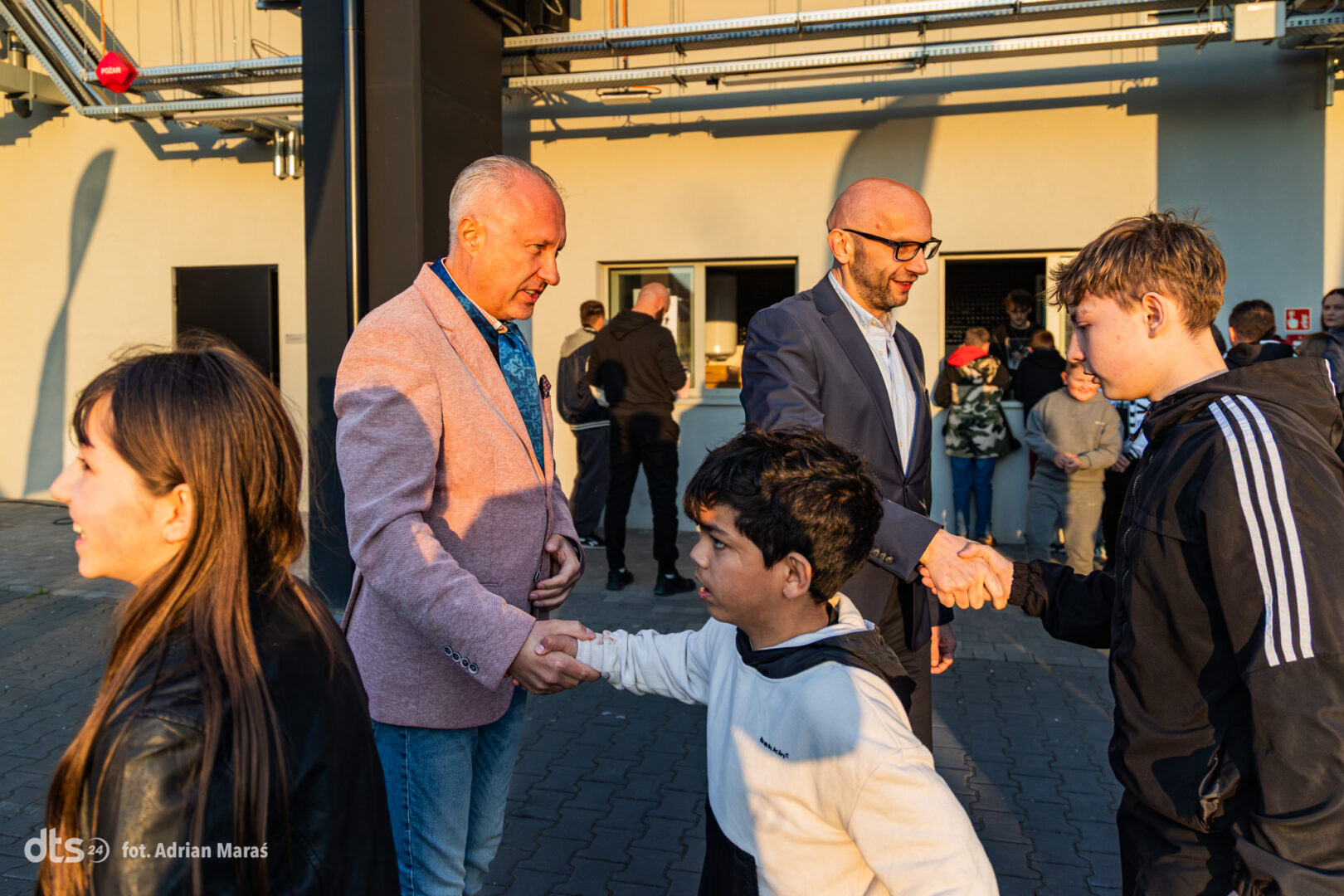 Two men in blazers shake hands with young people at an outdoor event, sunlit courtyard, others socializing in the background