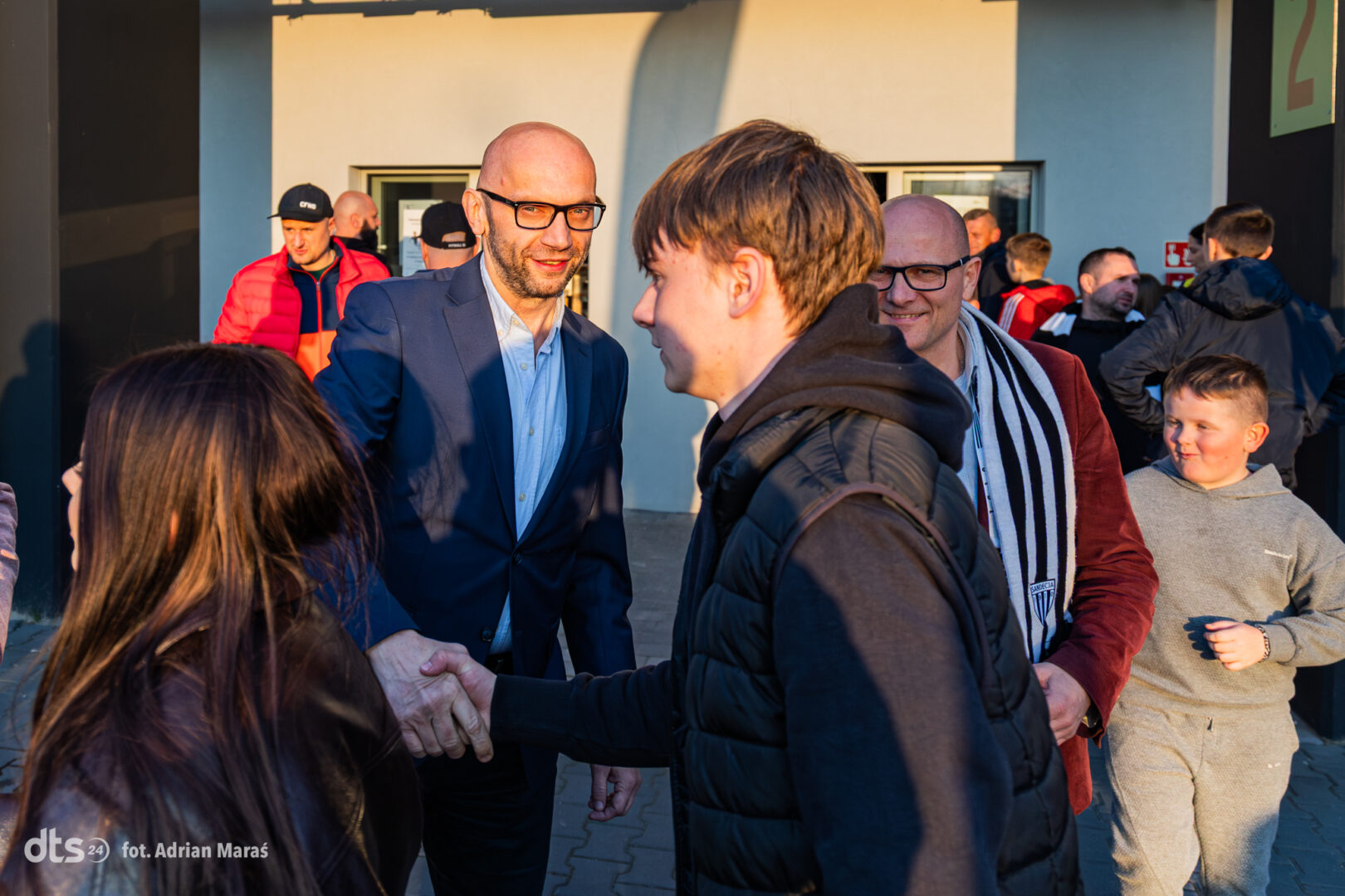 Man in a blue suit shakes hands with a younger man in a black vest while others chat nearby in an outdoor setting.