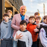 Group of people posing together outdoors near a modern building; man in a pink blazer stands with several children.