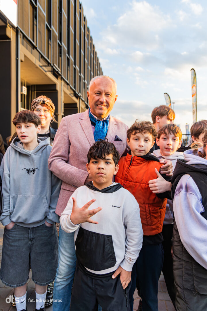 Group of people posing together outdoors near a modern building; man in a pink blazer stands with several children.