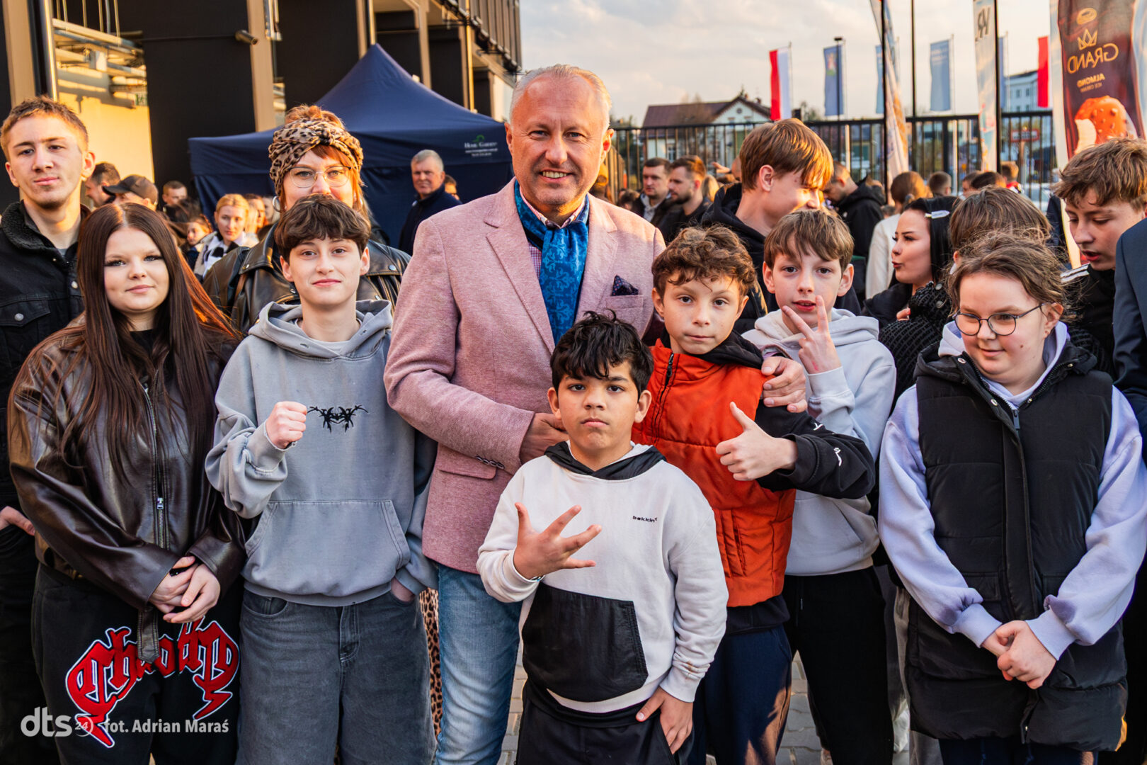 Group of smiling teens surrounding a man in a pink blazer at an outdoor event.