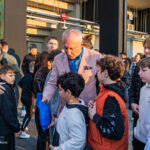 Older man in a pink blazer hands a blue drawstring bag to a boy among a group of children and adults outside a building.