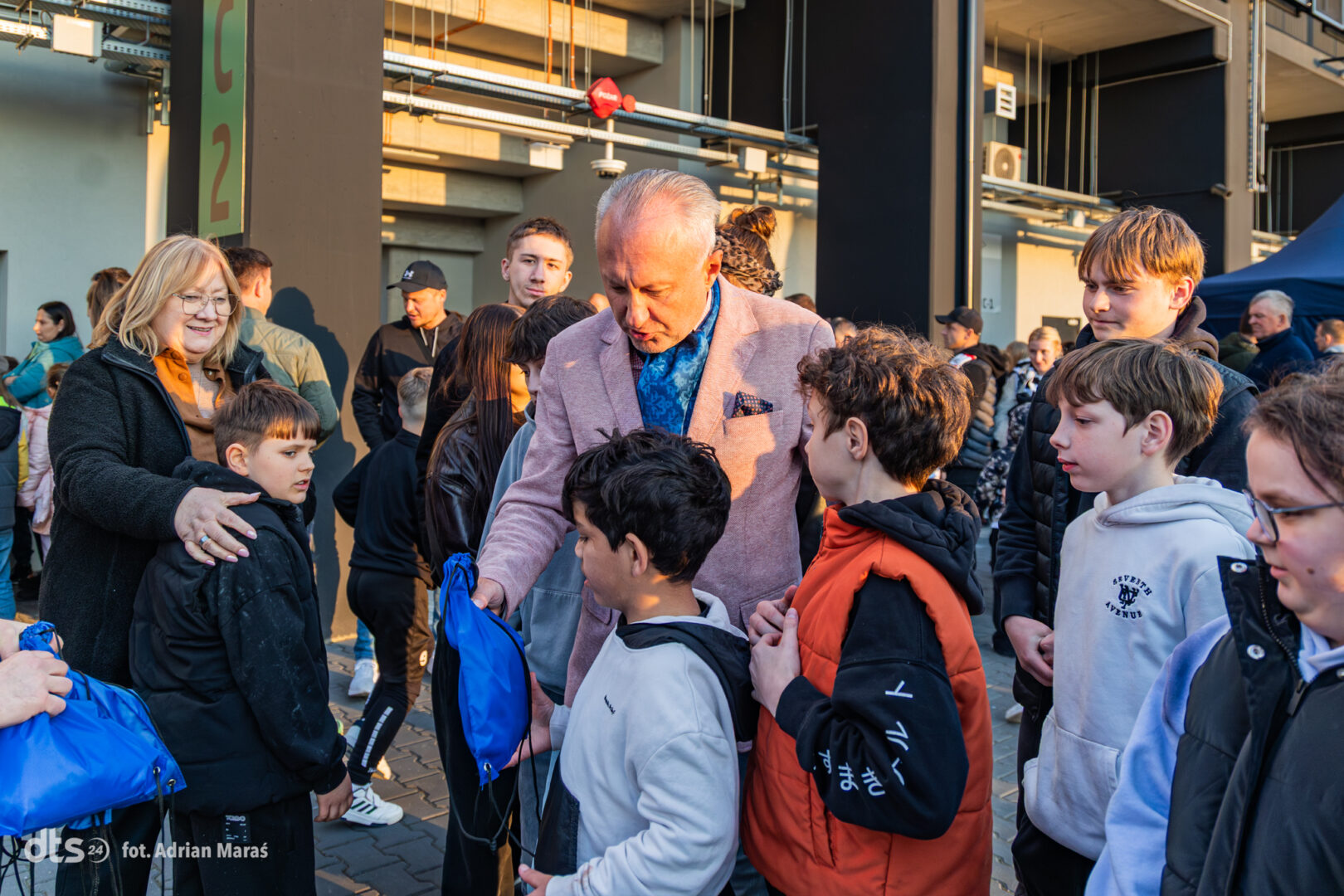Older man in a pink blazer hands a blue drawstring bag to a boy among a group of children and adults outside a building.