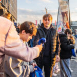 Teenager in a black puffer jacket accepts a blue bag from a person in a pink hoodie during an outdoor event, while others watch.