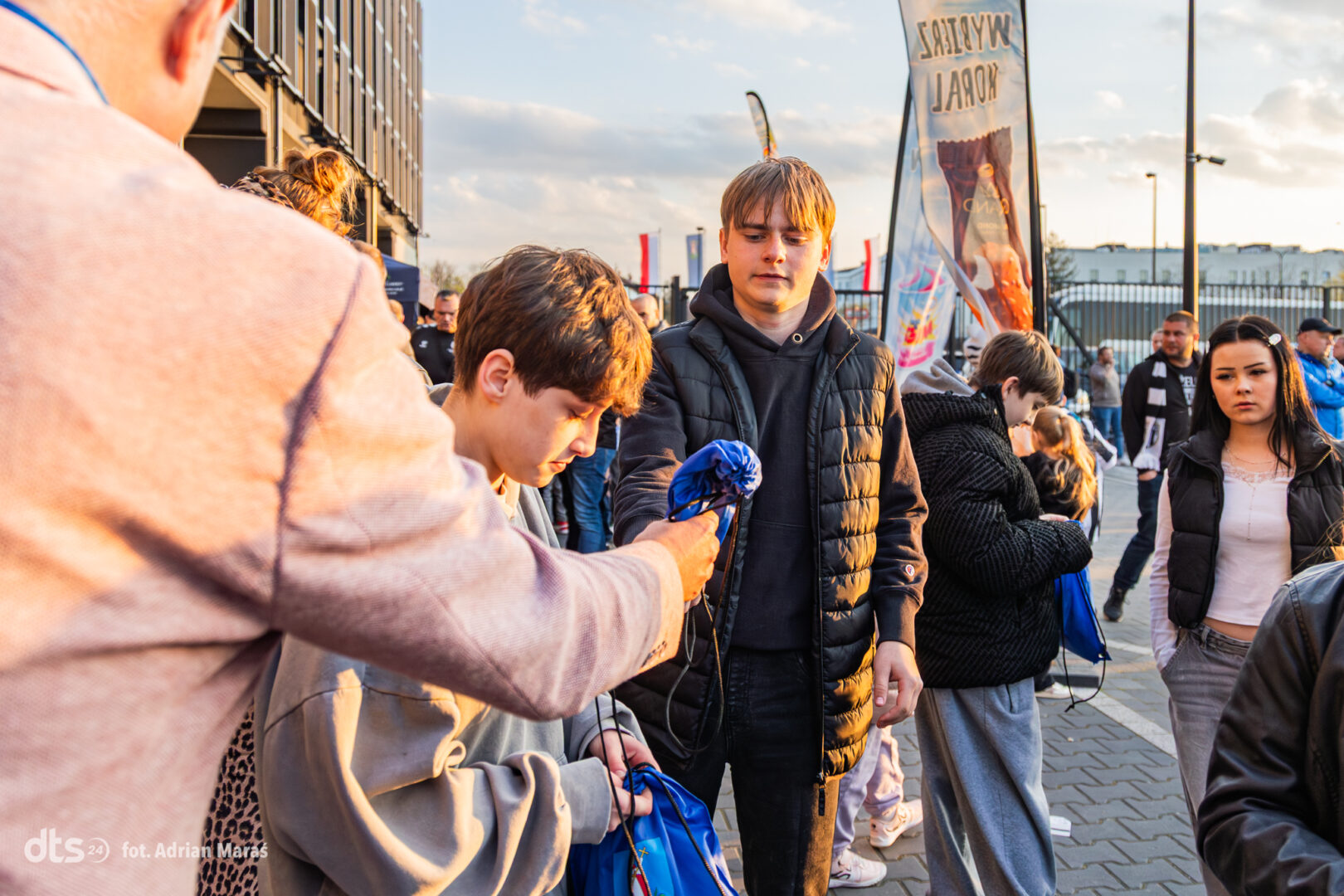 Teenager in a black puffer jacket accepts a blue bag from a person in a pink hoodie during an outdoor event, while others watch.