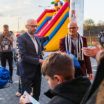 Man in a suit hands a blue bag to a child at an outdoor event near a colorful inflatable slide with a rainbow ramp backdrop.