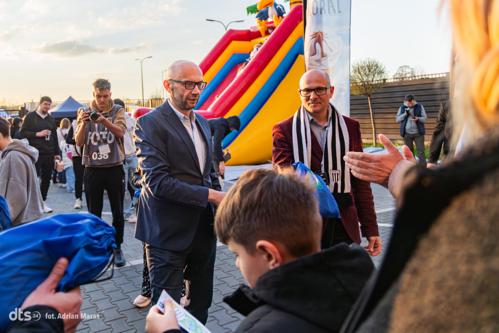 Man in a suit hands a blue bag to a child at an outdoor event near a colorful inflatable slide with a rainbow ramp backdrop.