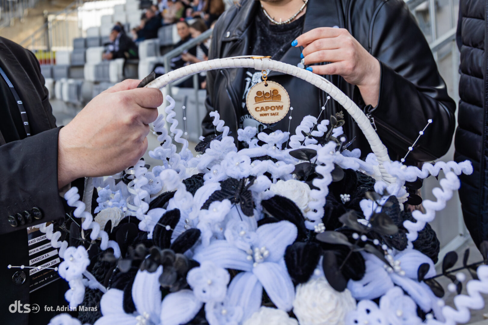 Hands shaping a white-and-blue floral basket, with a round CAPOW tag, at a stadium event. (informative)
