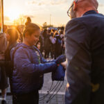 Boy in a blue puffer jacket hands a blue drawstring backpack to an adult at sunset during a crowded outdoor event.