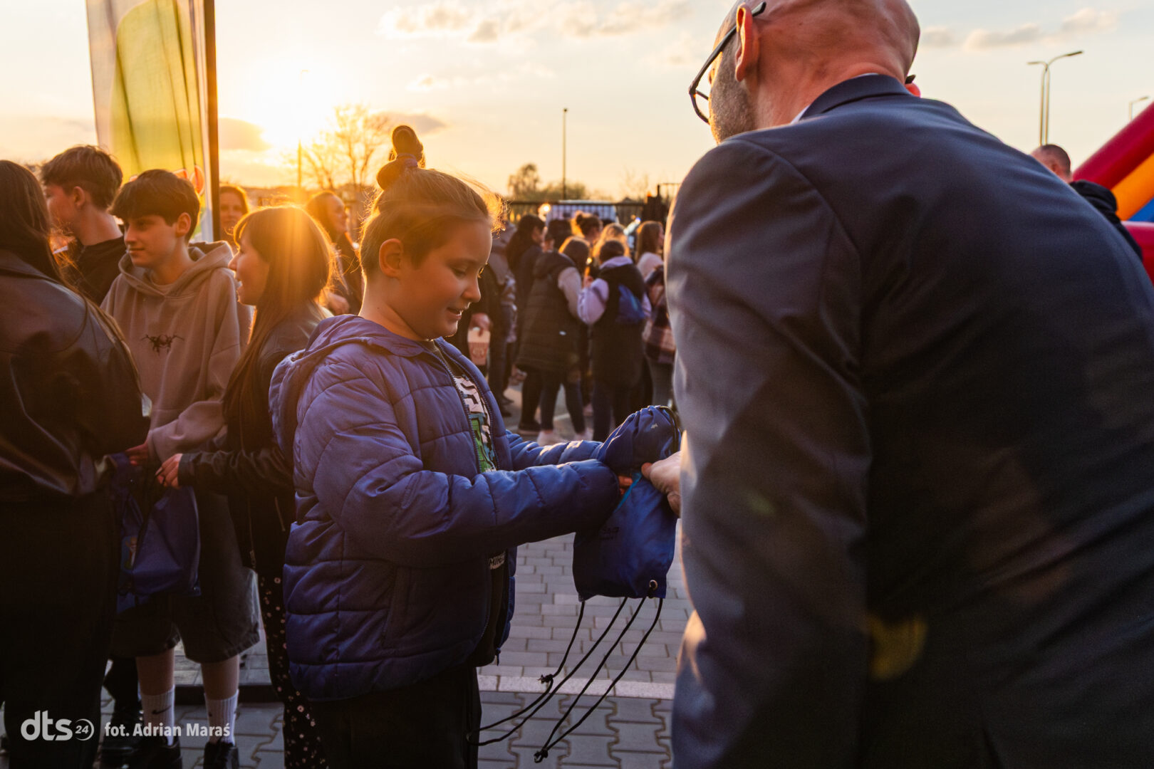 Boy in a blue puffer jacket hands a blue drawstring backpack to an adult at sunset during a crowded outdoor event.