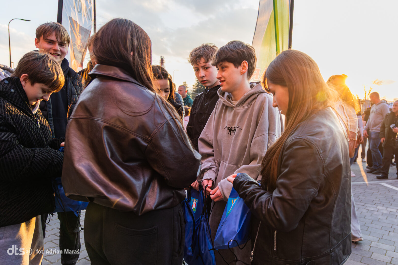 Group of teens standing in a circle outdoors at sunset, exchanging blue drawstring bags during an event.
