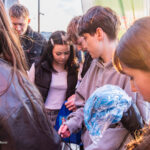 Group of teenagers outdoors, focusing on a blue bag being opened by a boy in a gray hoodie.