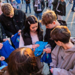 Group of teenagers gathering blue event bags from a distribution table in a sunny plaza.